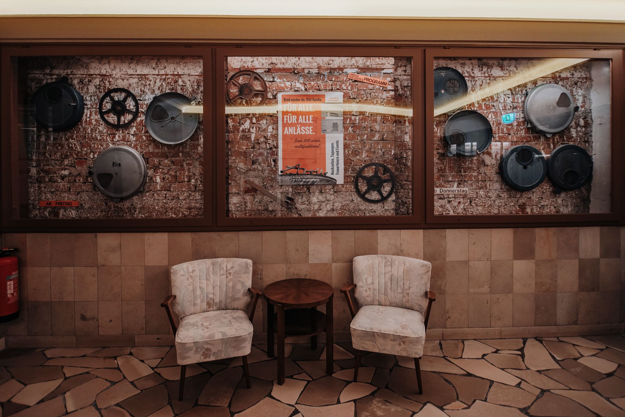 Zwei Retro-Sessel mit kleinem Holztisch vor einer Wand mit Filmrollen und Plakaten in einem nostalgischen Kinointerieur.