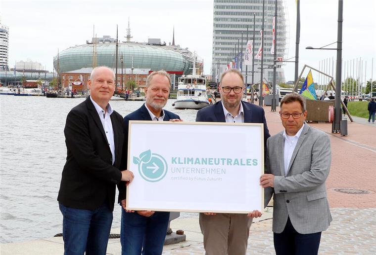 Vier Männer stehen an einem Hafen und halten ein Schild mit der Aufschrift „Klimaneutrales Unternehmen – certified by Fokus Zukunft“.