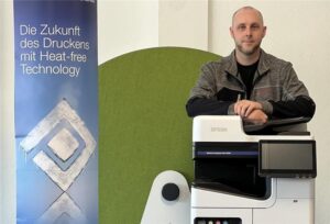 Mann steht neben einem modernen Epson-Drucker, daneben ein Banner mit der Aufschrift „Die Zukunft des Druckens mit Heat-free Technology“.