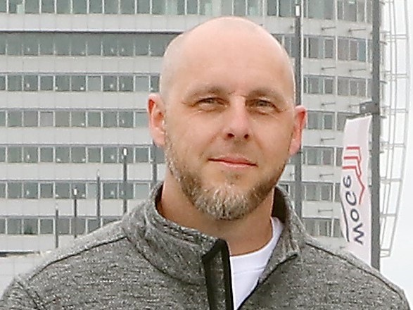 Porträt eines Mannes im grauen Pullover vor einem modernen Bürogebäude im Hintergrund.