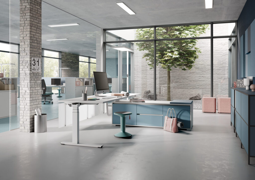 Helles Büro mit ergonomischem Steh-Sitz-Schreibtisch, Bildschirm, Stauraummöbeln und Blick durch Glaswände auf offene Arbeitsplätze und einen begrünten Innenhof