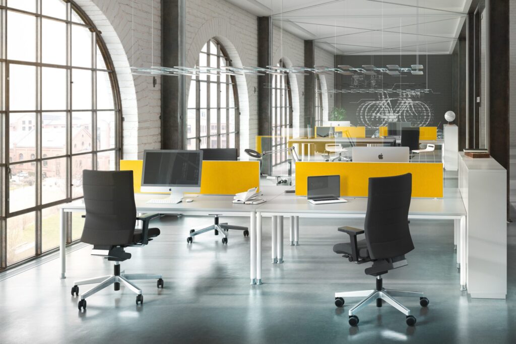 Modernes Großraumbüro mit weißen Schreibtischen, gelben Trennwänden, Computern und ergonomischen Bürostühlen vor großen Fensterbögen