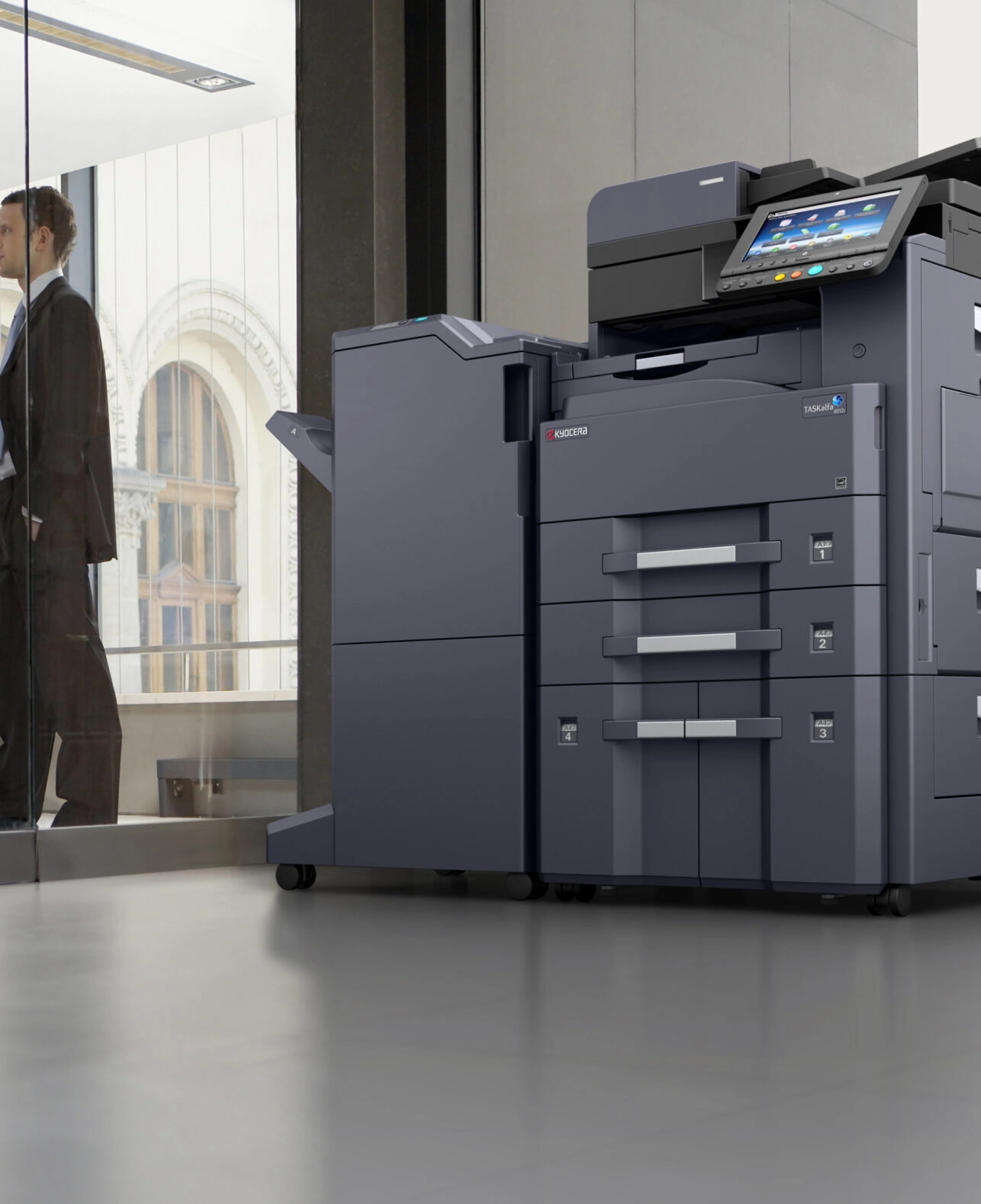 Großer grauer Kyocera-Multifunktionsdrucker in einem modernen Büro mit Glaswand und Geschäftspersonen im Hintergrund.