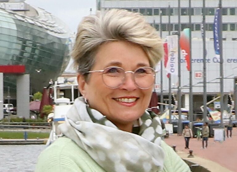 Frau mit Brille und graublondem Haar lächelt im Hafenbereich vor modernen Gebäuden.