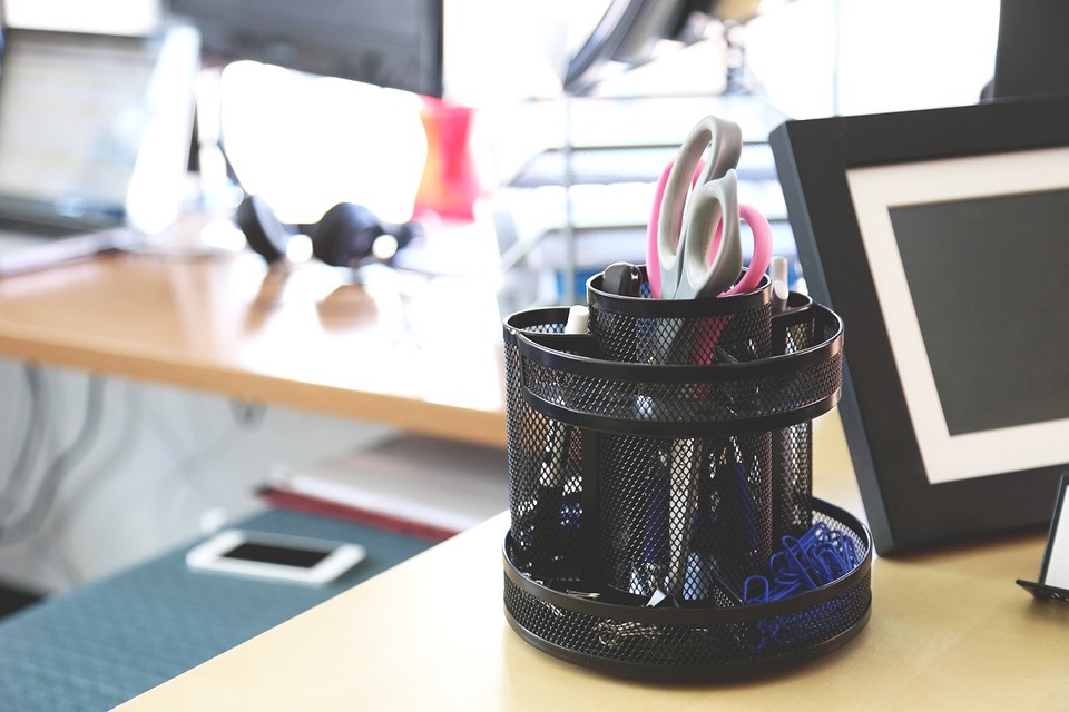 Schreibtisch mit schwarzem Stiftehalter, Scheren und Bürobedarf, im Hintergrund Laptop und Headset.