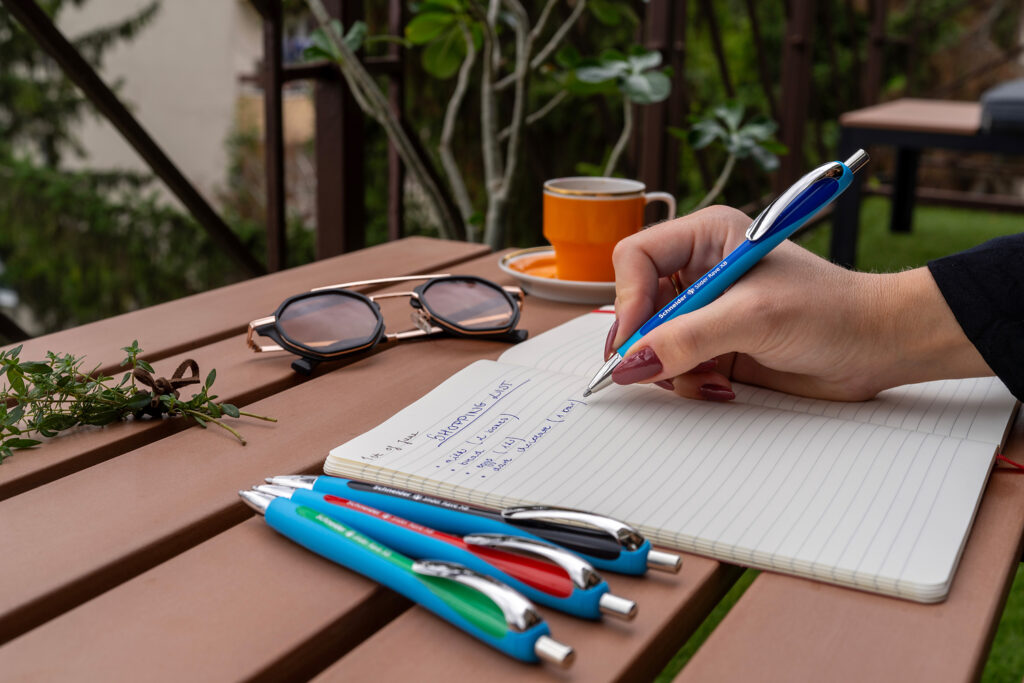 Hand schreibt mit blauem Kugelschreiber in ein Notizbuch auf einem Gartentisch, daneben liegen weitere Stifte, eine Sonnenbrille und eine Tasse Kaffee.