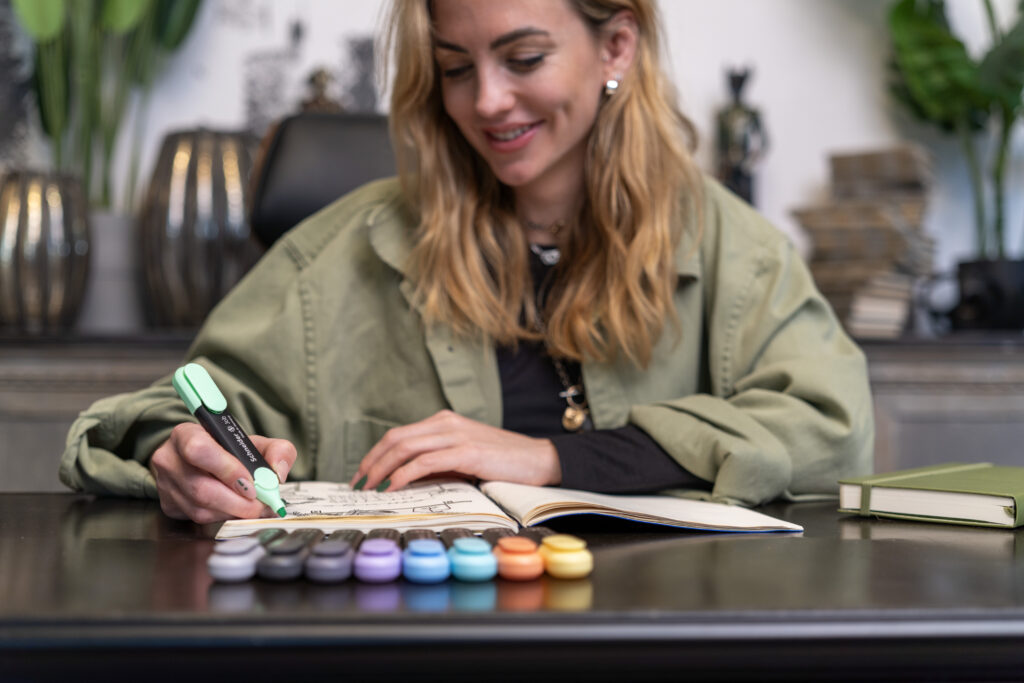 Frau markiert Text in einem Notizbuch mit einem grünen Textmarker, auf dem Tisch liegen mehrere bunte Marker – kreative Arbeitsszene im Büro.