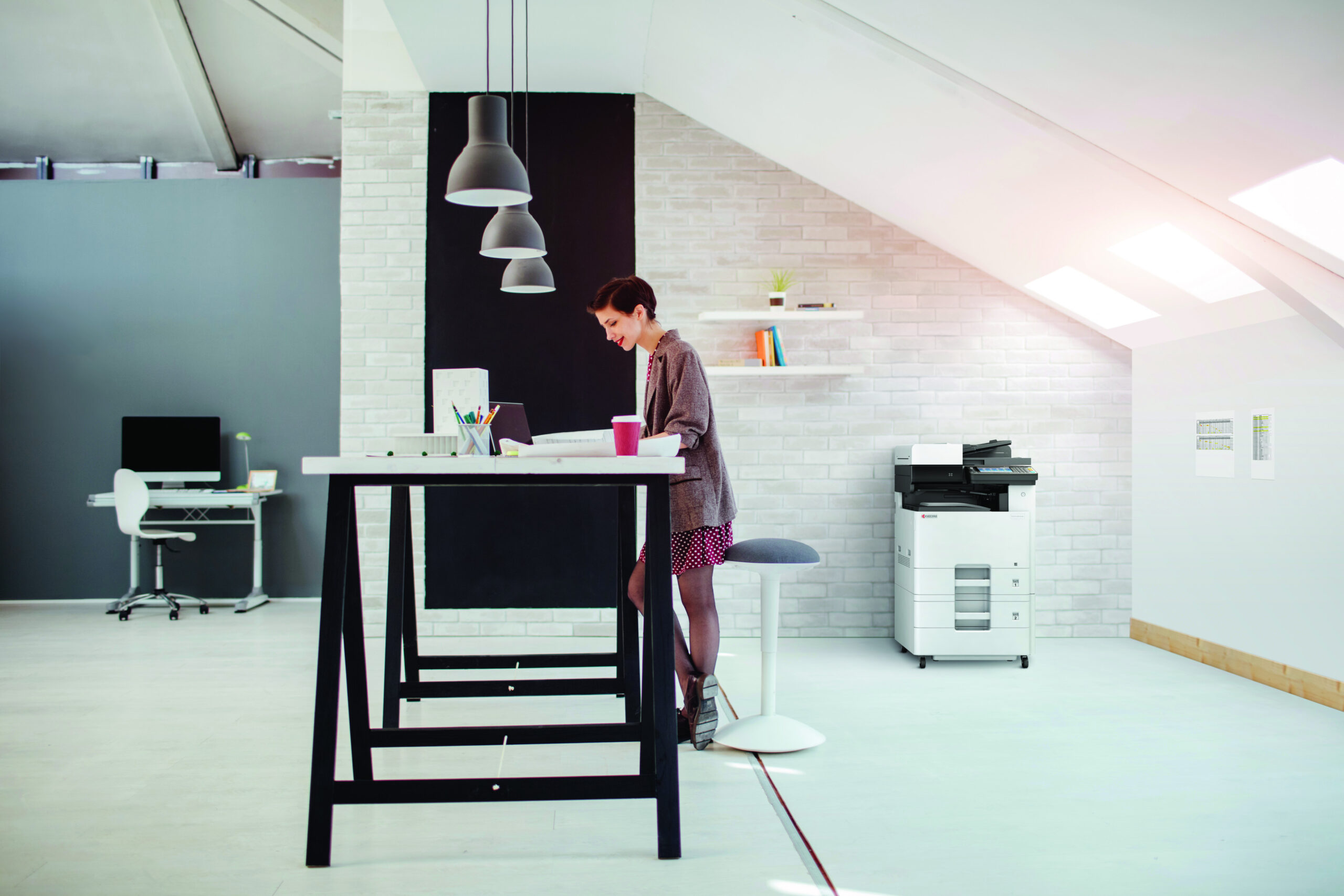 Frau arbeitet an einem hohen Schreibtisch in einem modernen, hellen Büro mit Kyocera-Drucker im Hintergrund.