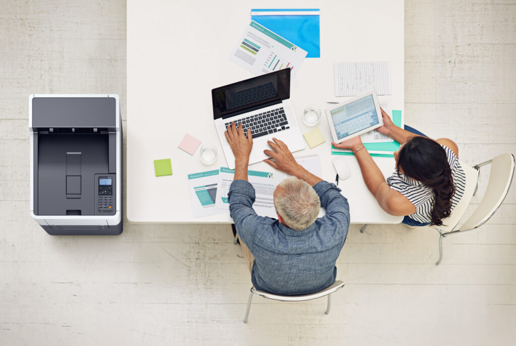 Zwei Personen arbeiten gemeinsam an einem Schreibtisch mit Laptop, Tablet und Dokumenten neben einem modernen Drucker.