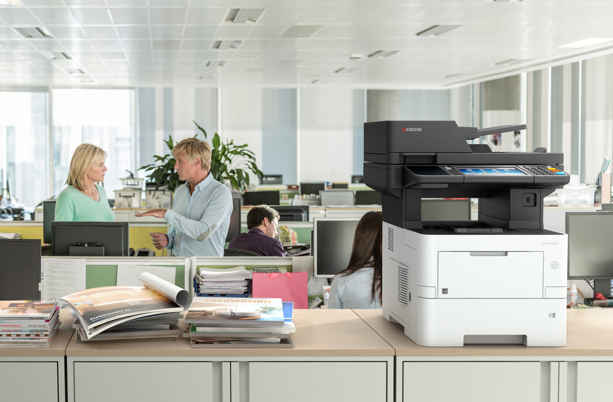 Offene Büroumgebung mit Mitarbeitern im Gespräch und einem großen Kyocera-Multifunktionsdrucker im Vordergrund.