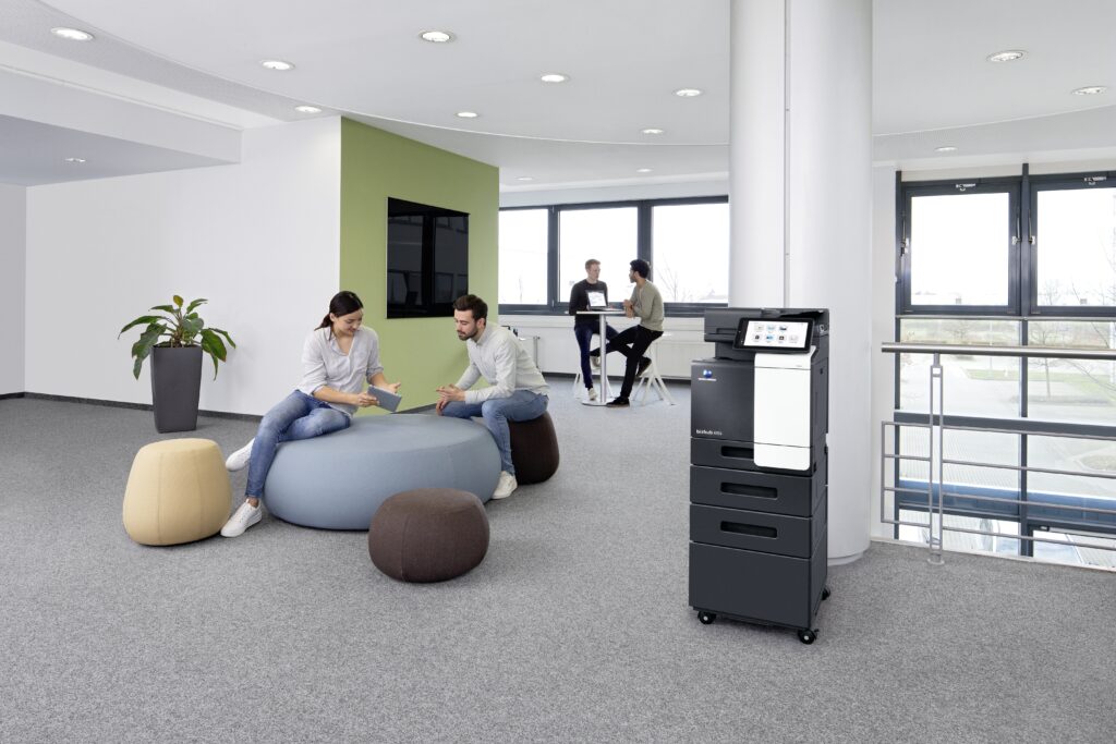 Helle Büroszene mit zwei Personen auf Sitzhockern im Gespräch, im Hintergrund ein Multifunktionsdrucker neben großen Fenstern.