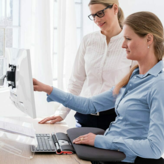 Zwei Frauen arbeiten gemeinsam an einem ergonomisch eingerichteten Arbeitsplatz mit höhenverstellbarem Monitor und Tastatur.