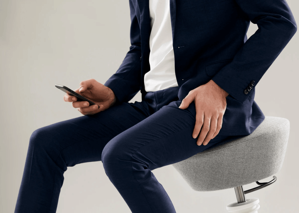 Mann in blauem Anzug sitzt auf ergonomischem Hocker und hält ein Smartphone in der Hand.
