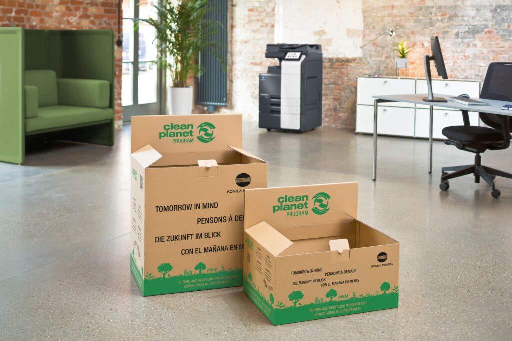 Zwei Recyclingboxen des „Clean Planet Program“ von Konica Minolta stehen in einem modernen Büro neben einem Multifunktionsdrucker.