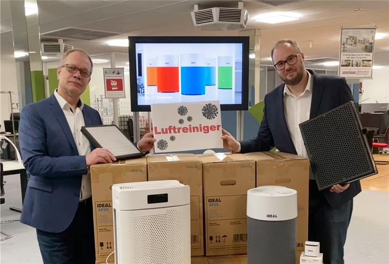 Zwei Männer präsentieren in einem Büro verschiedene Luftreiniger und Filterelemente vor gestapelten Verpackungskartons.
