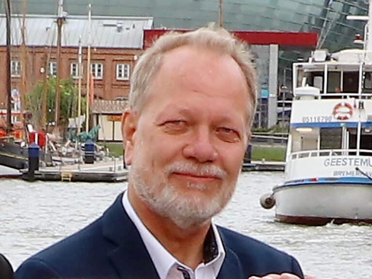 Mann mit grauem Haar, steht im Hafenbereich vor modernen Gebäuden und einem Schiff.