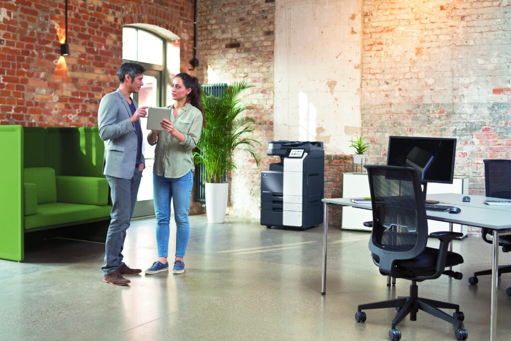 Zwei Personen im modernen Büro mit Backsteinwand besprechen etwas an einem Tablet, im Hintergrund steht ein Multifunktionsdrucker.