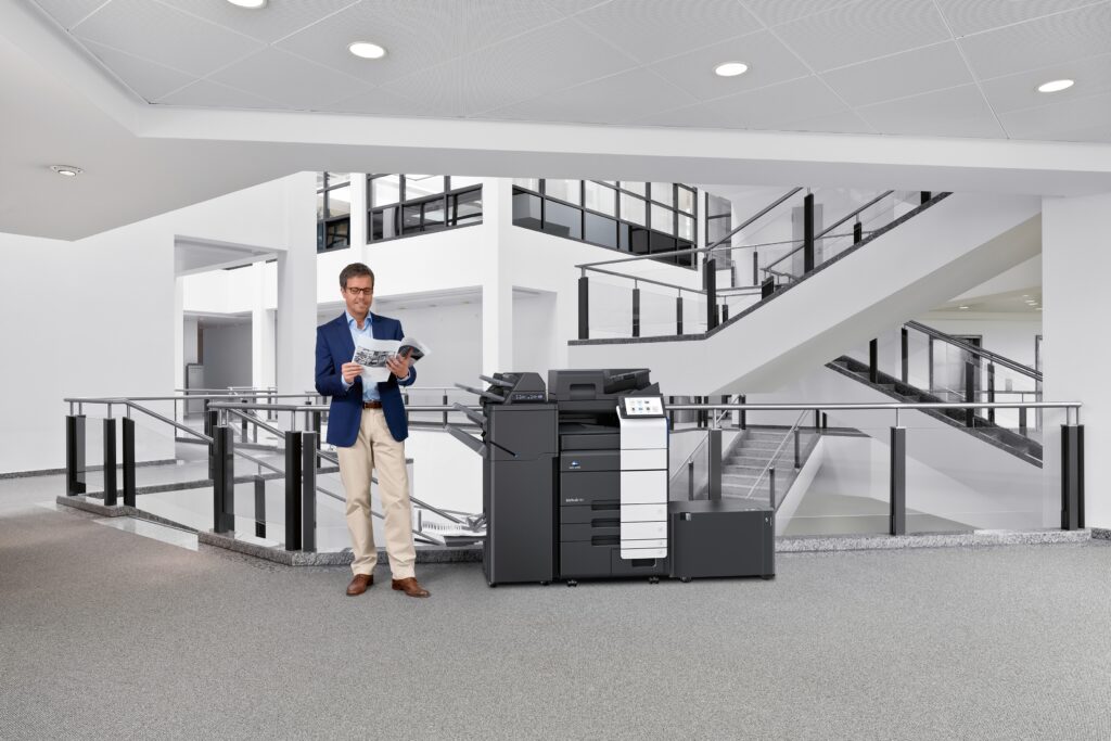 Mann im modernen Bürogebäude steht neben einem großen Multifunktionsdrucker und überprüft Ausdrucke.