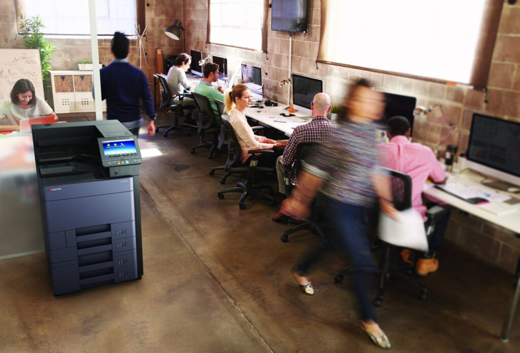 Modernes Großraumbüro mit mehreren arbeitenden Personen an Computern und einem großen Multifunktionsdrucker im Vordergrund.