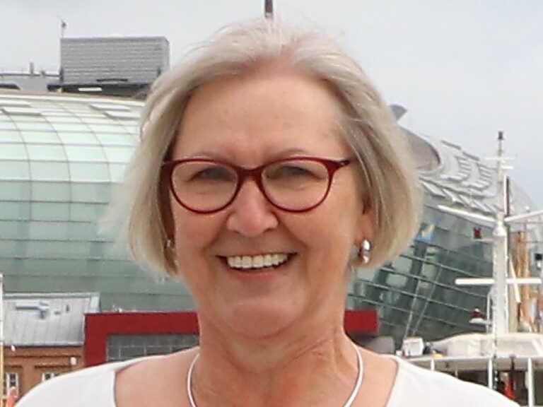 Porträt einer lächelnden Frau mit kurzem grauem Haar und roter Brille im Freien vor moderner Architektur.