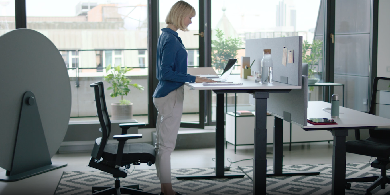 Frau arbeitet im Stehen an einem höhenverstellbaren Schreibtisch in modernem Büro mit Laptop und Pflanzen.