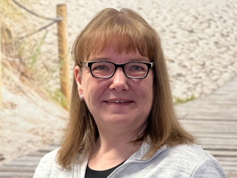 Porträt einer Frau mit Brille und hellbraunen Haaren vor einem Hintergrund mit Sandweg und Dünen.