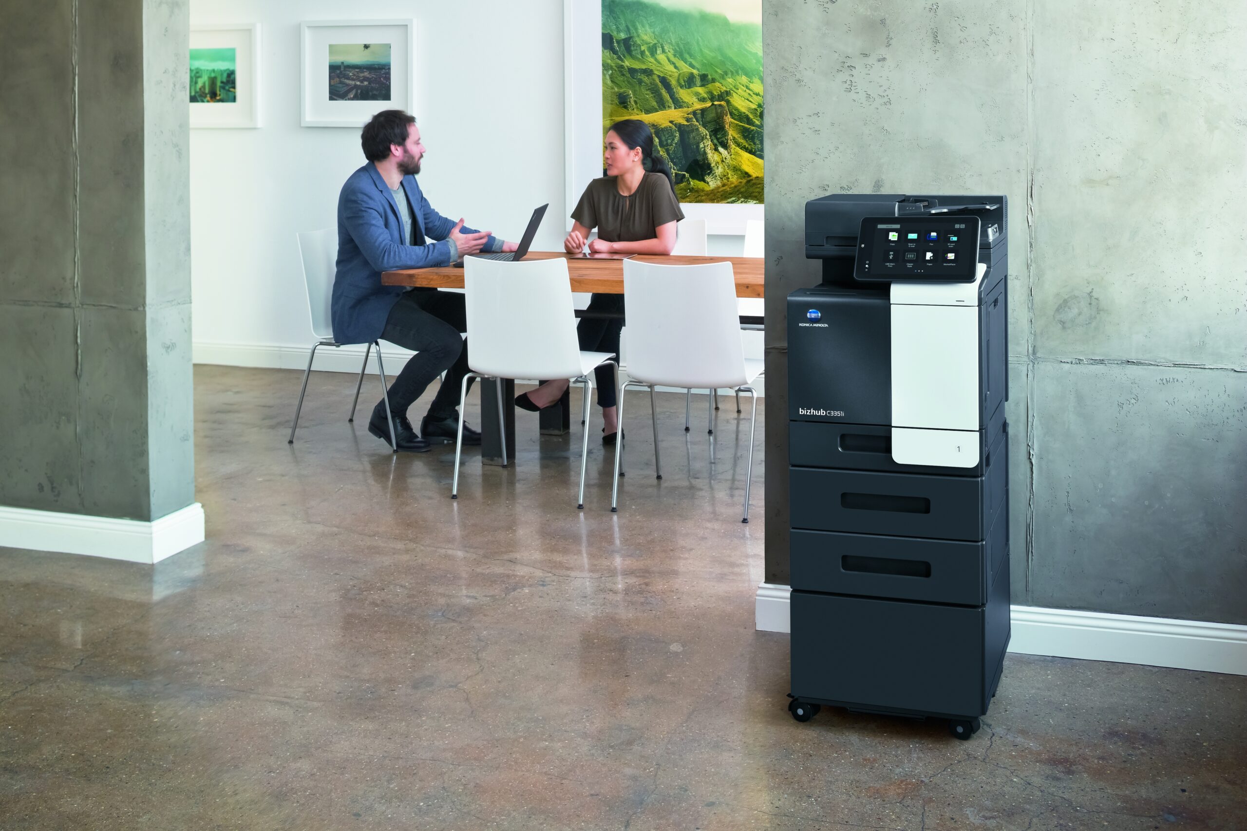 Modernes Büro mit zwei Personen im Gespräch an einem Holztisch und einem Multifunktionsdrucker im Vordergrund.