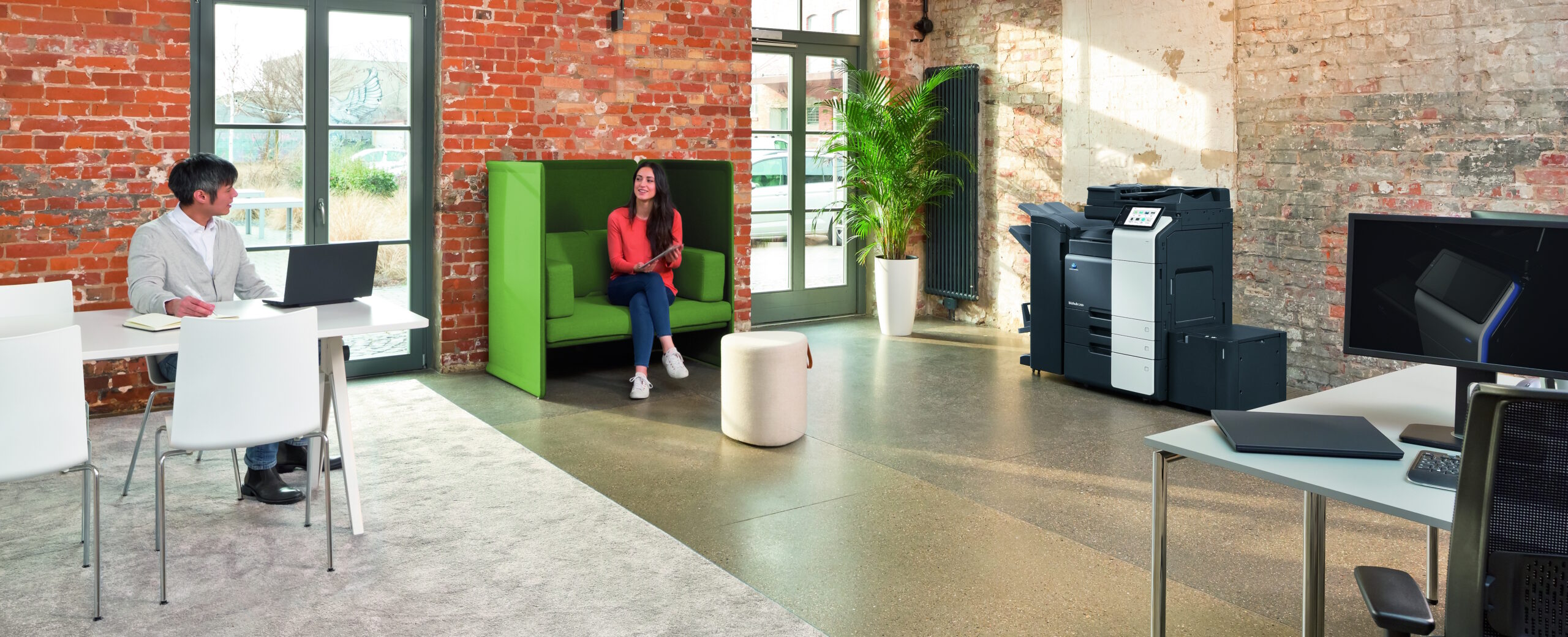 Modernes Büro mit Backsteinwänden, großer Multifunktionsdrucker, Mitarbeiter im Gespräch und grüner Sitznische.