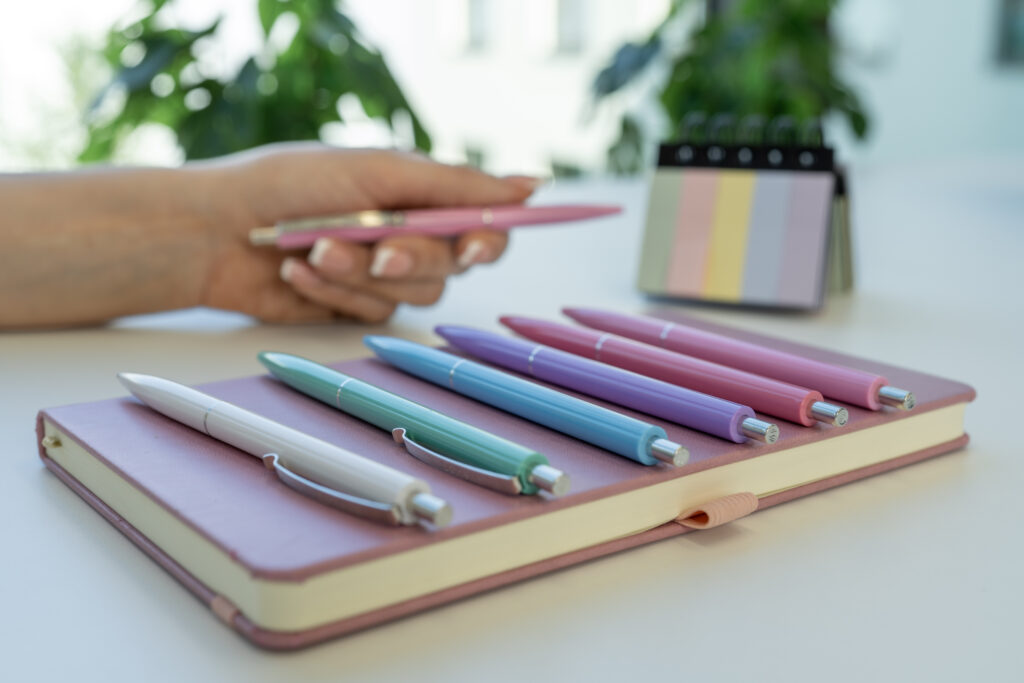 Mehrere bunte Kugelschreiber liegen ordentlich auf einem Notizbuch, im Hintergrund hält eine Hand einen Stift.