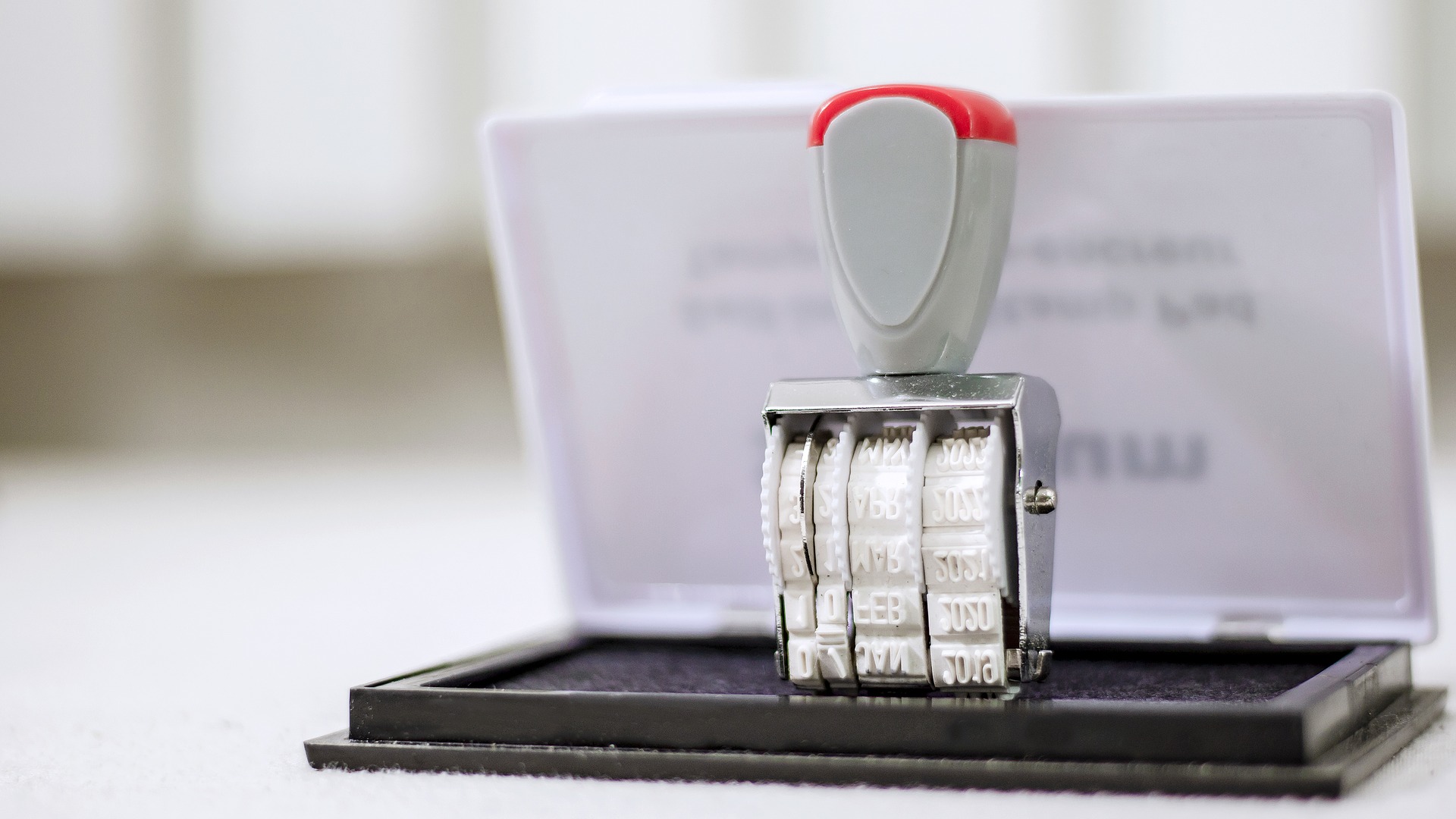 Datumsstempel mit rotem Griff steht auf einem schwarzen Stempelkissen vor hellem Hintergrund.