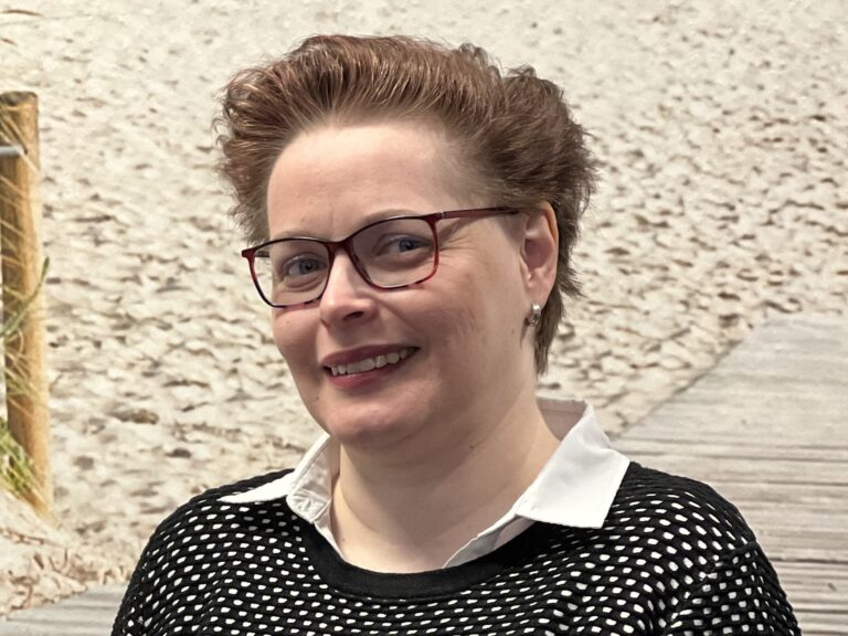 Porträt einer Frau mit Brille und braunen Haaren vor einem Hintergrundbild mit Sandweg und Dünen.