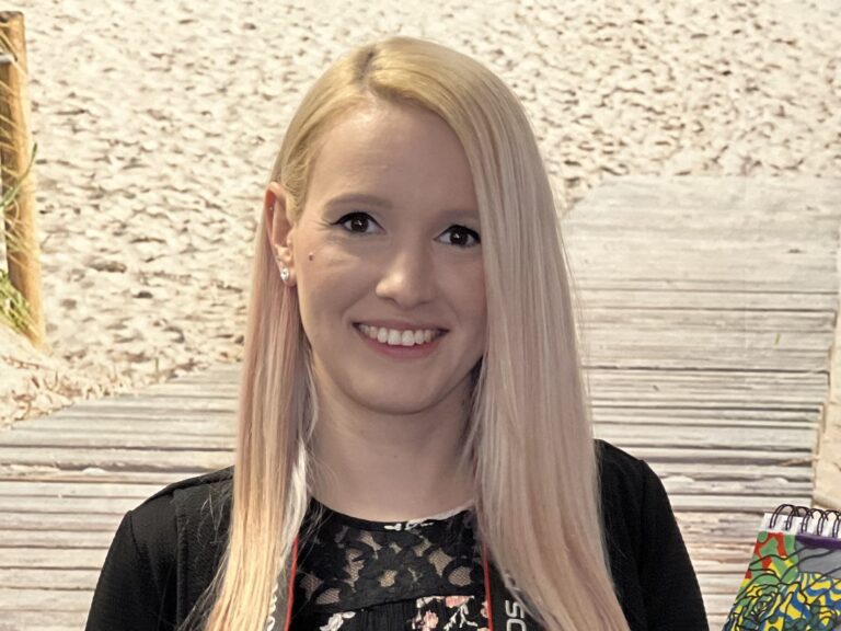 Porträt einer lächelnden Frau mit langen blonden Haaren vor einem Hintergrund mit Holzsteg und Sand.