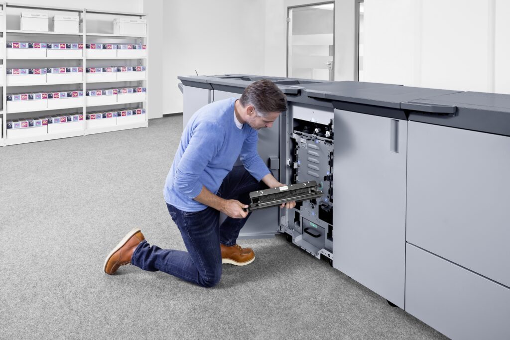 Mann kniet vor großem Drucksystem und führt Wartungsarbeiten an der geöffneten Maschine durch.