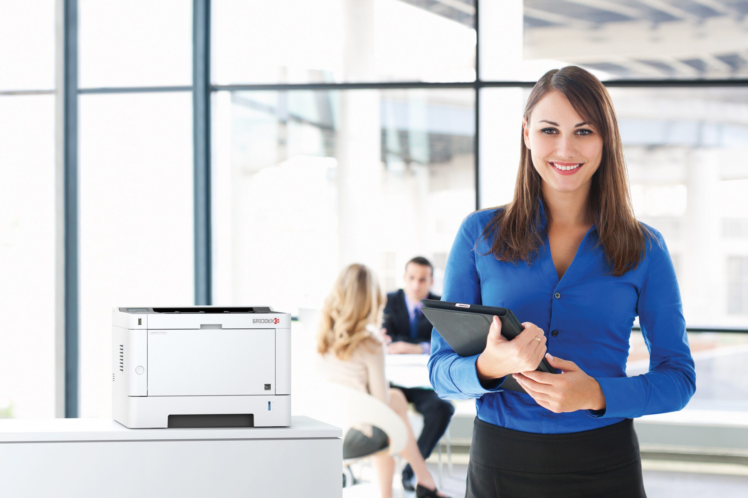 Geschäftsfrau mit Tablet steht lächelnd in modernem Büro neben einem weißen Kyocera-Drucker.