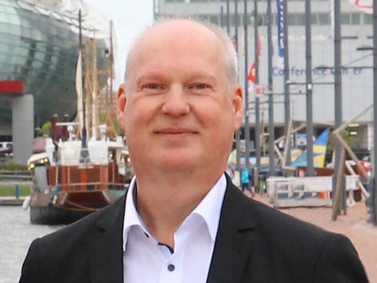 Mann mit grauem Haar, steht im Hafenbereich vor modernen Gebäuden.