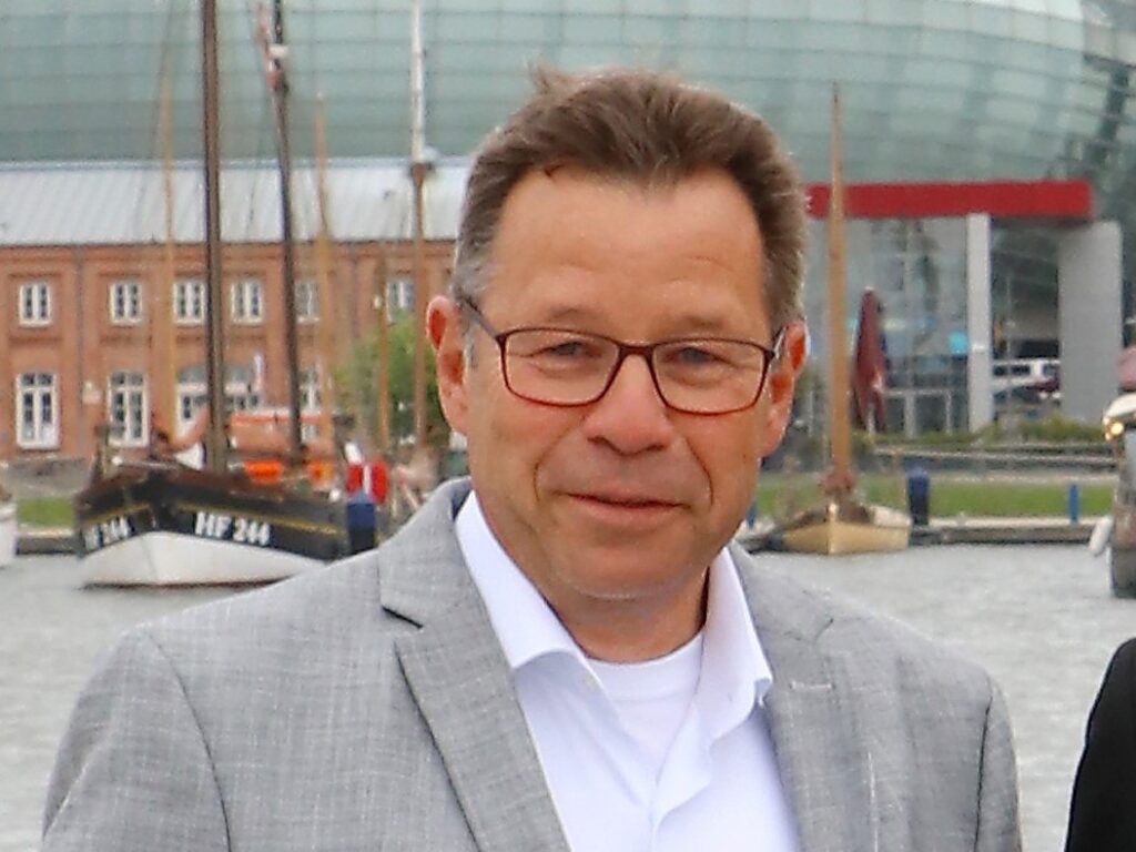 Mann mit Brille und grauem Sakko steht lächelnd im Hafenbereich, im Hintergrund sind Segelboote und Gebäude zu sehen.