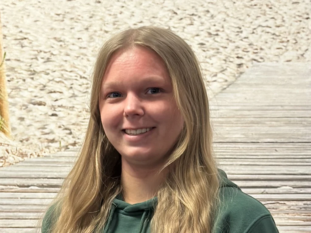 Frau mit langen blonden Haaren und grünem Hoodie lächelt in die Kamera, im Hintergrund ein Holzsteg am Sandstrand.