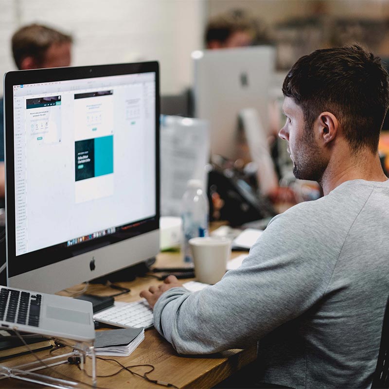 Mann arbeitet konzentriert am Computer in einem modernen Büro mit mehreren Monitoren und Schreibtischen.